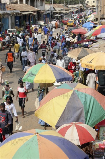 GAMBIA, Western Gambia, Serekunda, "Bakau Market, Atlantic Road.  Busy city street lined with stalls beneath colourful striped sun umbrellas.  Crowds of people, traffic and shop fronts open to the street.                   "