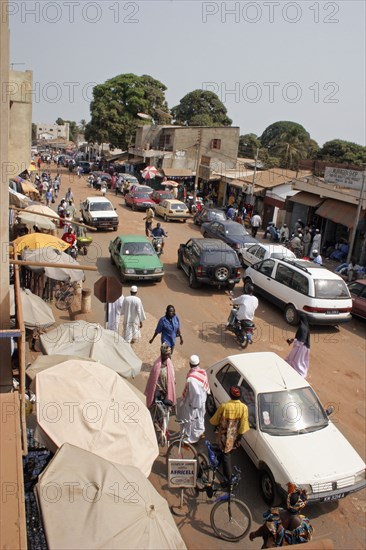 GAMBIA, Western Gambia, Serekunda, "Bakau Market,  Atlantic Road.  Main city road lined with street stalls beneath sun umbrellas and open fronted shops with people and cars. "