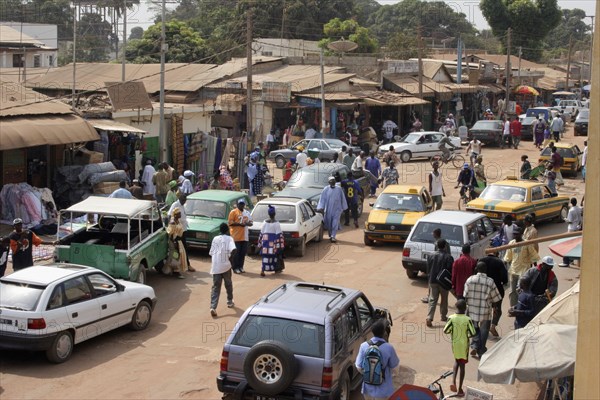 GAMBIA, Western Gambia, Serekunda, "Bakau Market, Atlantic Road.  View of Serekunda's main city street lined with open fronted shops and roadside stalls beneath sun umbrellas, people and vehicles. "