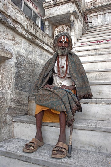 INDIA, Rajasthan, Udaipur, Elderly male Hindu beggar sitting on steps outside the Jagdish Temple wrapped in blanket shawl with painted forehead and grey beard.