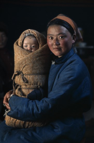 MONGOLIA, People, Young mother in fleece-lined silk tunic holding baby wrapped in traditional swaddling against bitter winter cold. East Asia Asian Babies Classic Classical Historical Kids Mongol Uls Mongolian Mum Older