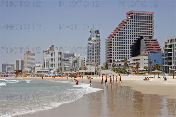 ISRAEL, Tel Aviv, "Beach with people sunbathing and at waters edge overlooked by high rise, modern city buildings reflected on wet sand."