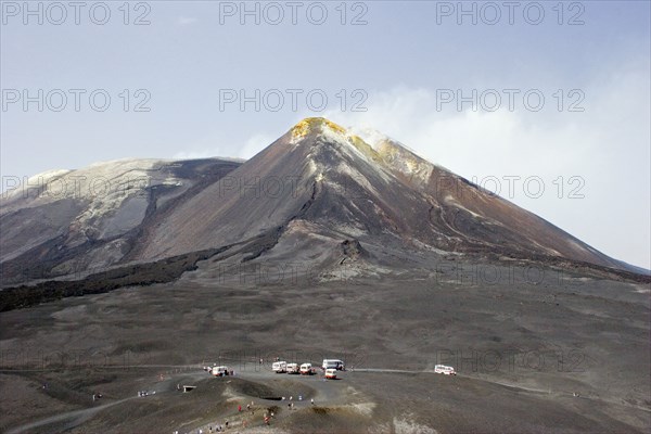 ITALY, Sicily, Mount Etna, View of active and smoking main crater of volcano and older crater with visitors and transportation vehicles in foreground.