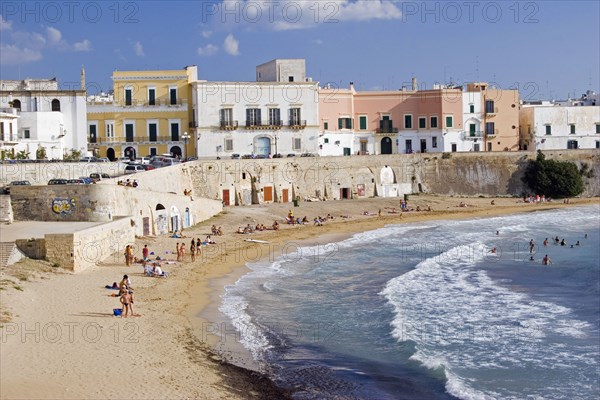ITALY, Puglia, Gallipoli, View of old city with traditional houses beside sea wall overlooking curving sandy beach with people sunbathing and swimming