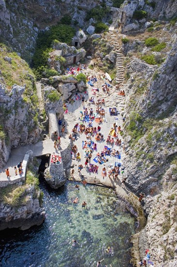 ITALY, Puglia, Lecce, Tricase.  Looking down on people sunbathing on coloured beach towels laid out on rocks next to the sea with others in the water.  Steps leading down behind and cliff path at side.
