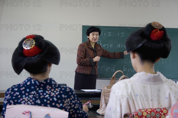 JAPAN, Honshu, Kyoto, "Gion area the neighbourhood where Geisha live, study and perform.  Two Geisha wearing kimono and wigs attend an English class at Mia Garatso school for Geisha, seated with backs to camera looking towrds teacher pointing to blackboard."