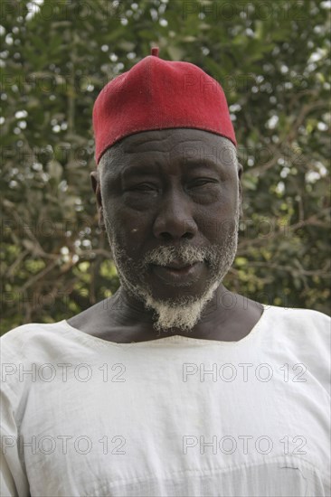 GAMBIA, Western Gambia, Tanji, Tanji Village.  Head and shoulders portrait of Muslim man with white trimmed beard wearing white shirt and red cap.