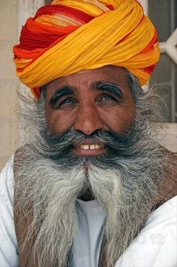 INDIA, Rajasthan, Jodhpur, "Meherangarh Fort.  Head and shoulders portrait of a smiling Indian man with long, grey beard wearing red, yellow and orange turban."