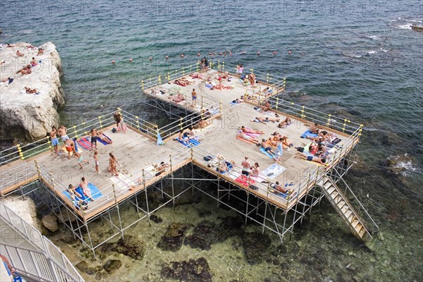 ITALY, Sicily, Syracuse, "Looking down on platform structure raised above rocky sea with steps to water, people sunbathing, playing and swimming."