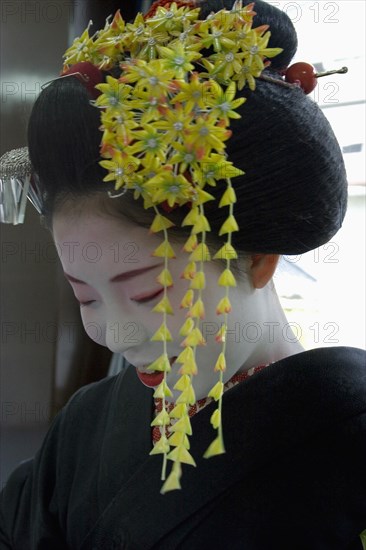 JAPAN, Honshu, Kyoto, Gion District.  Portrait of a Maiko apprentice Geisha with white painted face and red painted lips wearing black kimono and hair worn up with decorative pins and flower ornamentation ready for performance.