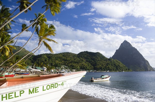 WEST INDIES, St Lucia, Soufriere, Fishing boats on the beach lined with coconut palm trees with the town and the volcanic plug mountain of Petit Piton beyond. A fishing boat in the foreground with the words Help Me Lord written on the bow