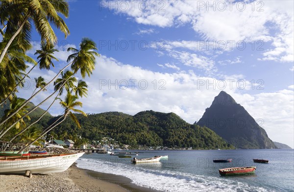 WEST INDIES, St Lucia, Soufriere, Fishing boats on the beach lined with coconut palm trees with the town and the volcanic plug mountain of Petit Piton beyond. A fishing boat in the foreground with the words Help Me Lord written on the bow