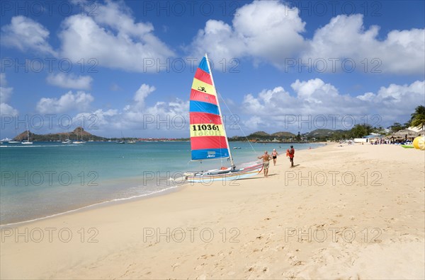 WEST INDIES, St Lucia, Gros Islet, Reduit Beach in Rodney Bay with tourists on the beach beside a hobbycat catamarran with yachts at anchor in the bay and Pigeon Island in the distance