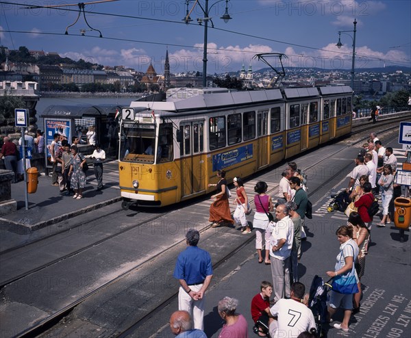 HUNGARY, Budapest, Electric yellow tram and groups of people gathered at tram stop