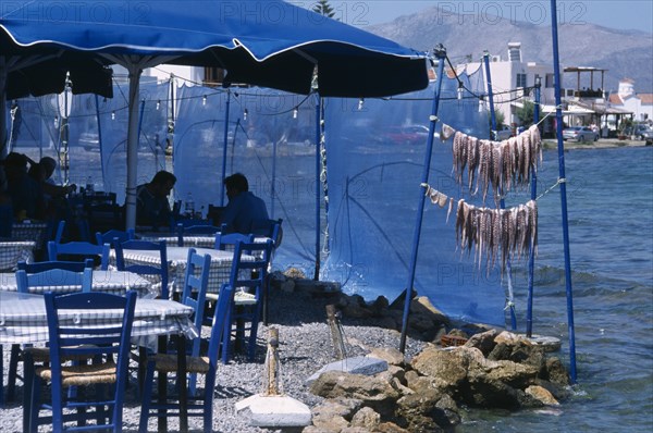 GREECE, Peloponnese, Elafonisos , Restaurant outside seating with blue table and chairs next to waters edge with a fresh catch of seafood hanging from poles