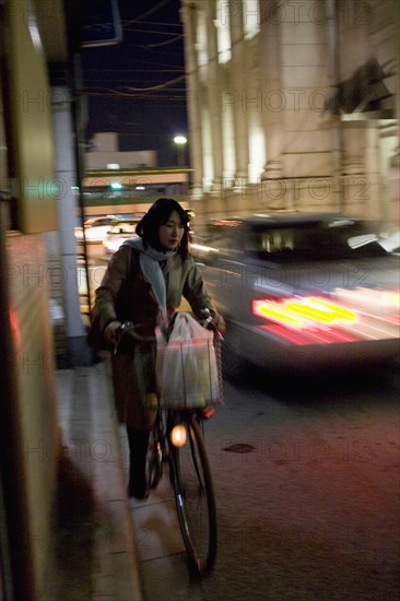 JAPAN, Honshu, Kyoto, Gion District.  Japanese woman riding her bicycle at night along a side road in Kyoto carrying bag of shopping in basket on the handlebars with car passing in the opposite direction.
