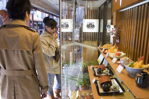 JAPAN, Honshu, Tokyo, Asakusa.  Young Japanese couple standing outside a restaurant looking at the plastic food representing the menu displayed in the window