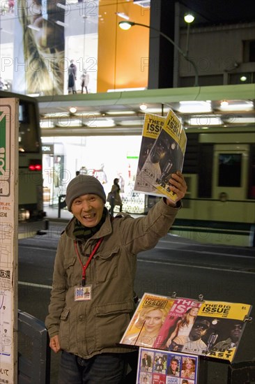 JAPAN, Honshu, Kyoto, "Japanese man selling the ‘Big Issue’ magazine, arm raised, holding copy with passing traffic on street behind and lighted shop windows in evening."
