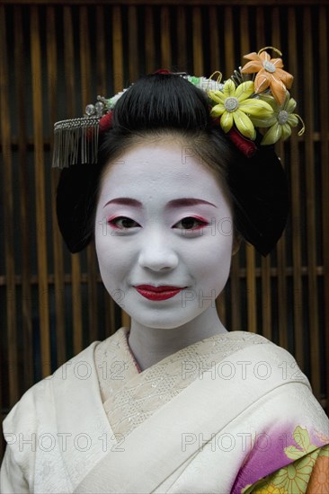 JAPAN, Honshu, Kyoto, "Gion District.  Head and shoulders portrait of smiling apprentice Geisha or Maiko with hair pinned up with flowers and decorative ornaments, white facial  make up, red painted lips, and wearing traditional kimono"