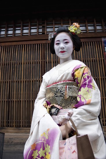 JAPAN, Honshu, Kyoto, "Gion District, the neighbourhood where Geisha live and perform.  Three-quarter portrait of smiling Maiko, or apprentice Geisha with hair pinned up with decorative ornaments, white facial make up, red painted lips and wearing traditional kimono of pale, flower patterned silk. "