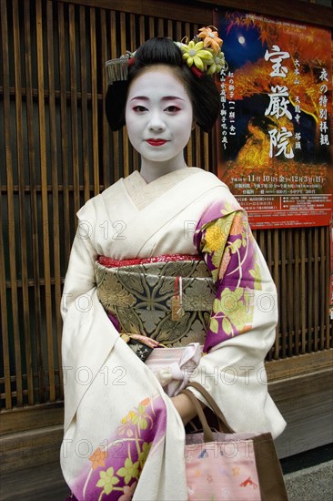 JAPAN, Honshu, Kyoto, "Gion District.  Three-quarter standing portrait of Geisha with white facial make up and red painted lips, hair worn up with decorative pins and wearing flower patterned, pale silk kimono. "