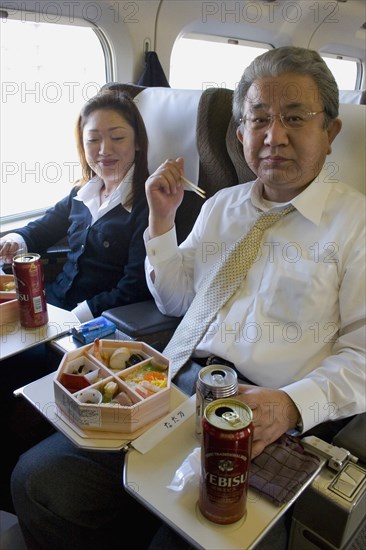 JAPAN, Honshu, Shinkansen train series 700 known as the ‘bullet train’.  Japanese couple eating from bento lunchbox using chopsticks while travelling between Tokyo and Kyoto.