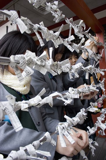 JAPAN, Honshu, Tokyo, Asakusa Kannon or Senso-ji Temple.  Three young Japanese female students hanging their omikuji folded fortune telling paper slips on bars outside the temple.
