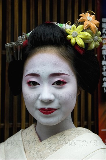 JAPAN, Honshu, Kyoto, "Gion District.  Head and shoulders portrait of smiling Maiko or apprentice Geisha, with hair worn up and fixed with decorative pins and flower ornaments, white facial make up and red painted lips wearing kimono, part seen."