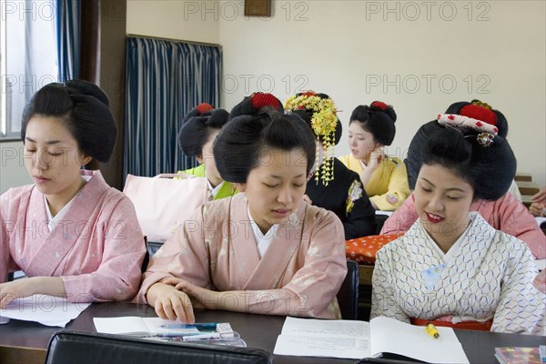JAPAN, Honshu, Kyoto, Gion District.  Geisha and Maiko apprentice Geisha attending a class at Mia Garatso school of Geisha.  Three young women wearing plain kimono seated at desk in foreground with open books.
