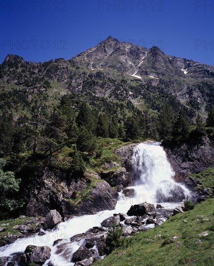 FRANCE, Midi-Pyrenees, Hautes-Pyrenees, Vallee du Lutour.  Jagged peaks of Soum det Guingays 2451 m / 8027 ft above white waterfall.  Outcrops of rock scattered over hillside amongst trees.