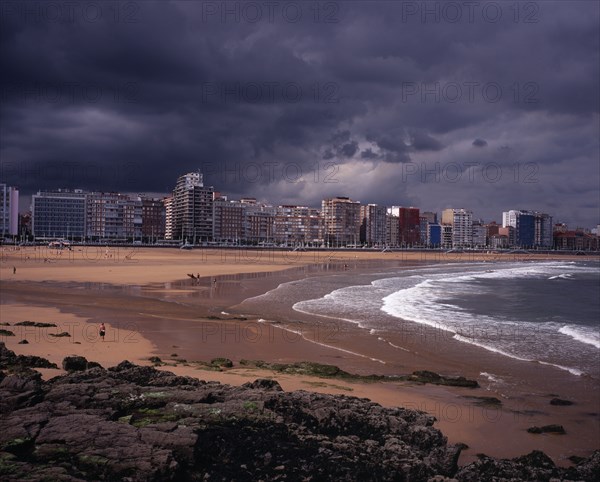 SPAIN, Asturias, Gijon, "High rise city buildings overlooking beach with people in water, on beach and surfing.  Grey, cloudy sky with approaching storm.  Rocks and seaweed in foreground."