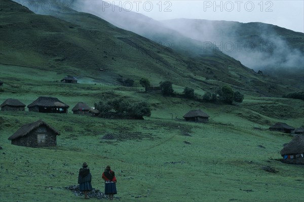 PERU, Ayacucho Province, Choquetira , Village of thatched homes on green hillside with mist seen high above. Two women standing together at the bottom of the slope