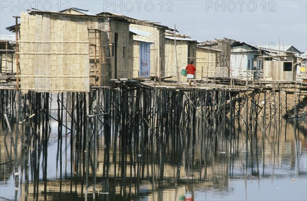 ECUADOR, Guayas Province, Guayaquil , Slum housing with stilt buildings built over untreated sewage. A woman wearing a red cardigan carrying a bucket along walkway with reflections on the water below