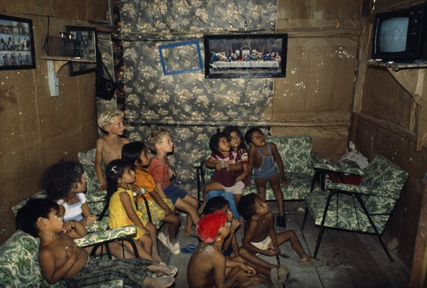 ECUADOR, Guayas Province, Guayaquil , Titus and Nathaniel Moser sitting with children watching television in Rosa’s house inside a Guayaquil slum. Framed painting of The Last Supper hanging on wall above the children.