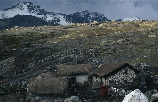 BOLIVIA, Northern Andes, Antaquilla , Collpa Huata Llama herders settlement of stone houses with thatched roofs. A woman wearing traditional red textiles standing next to stone wall. Snow covered mountains behind. Near Peru