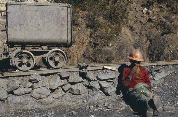 BOLIVIA, Chukiuta, Miner wearing orange hard hat sitting next to mining rail track chewing leaves of coca