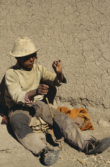 BOLIVIA, Chukiuta, Aymara man sitting against a wall with a spindle of thread. Near Sucre