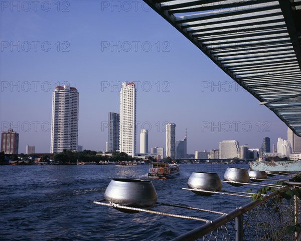 THAILAND, Bangkok, "Chao Phraya River from Wat Ratchasungkhon decorated with silver alms bowls, high rise buildings on opposite bank including Shangri-La hotel and new apartments with river ferry departing north for Saphan Thaksin bridge stop."