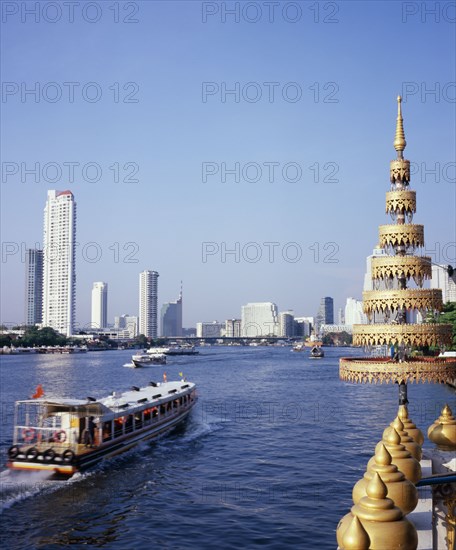 THAILAND, Bangkok, "View across Chao Phraya River from Wat Ratchasungkhon decorated with gold umbrella usually sheltering Buddha in foreground with express river ferry, indicated by orange flag departing north for Saphan Thaksin bridge stop.  New apartments, Shangri-la Hotel and high rise buildings on opposite bank."