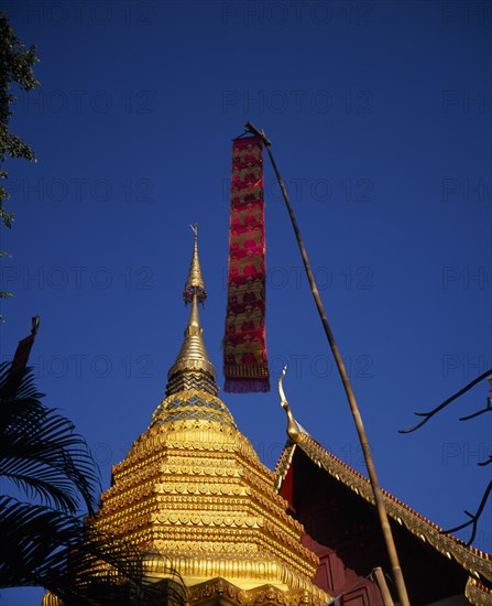 THAILAND, North, Chiang Mai, Wat Phran Tao.  Detail of pointed roof top of rare teak temple beside golden Chedi with red and gold prayer flag hanging in foreground.