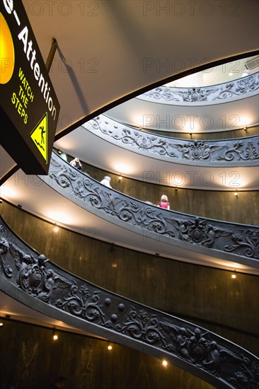 ITALY, Lazio, Rome, Vatican City Museums Tourists descending the Spiral Ramp designed by Giuseppe Momo in 1932 leading from the museums to the street level below seen from below with an illuminated sign warning to watch the steps