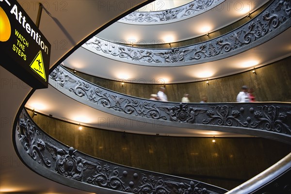 ITALY, Lazio, Rome, Vatican City Museums Tourists descending the Spiral Ramp designed by Giuseppe Momo in 1932 leading from the museums to the street level below seen from below with an illuminated sign warning to watch the steps