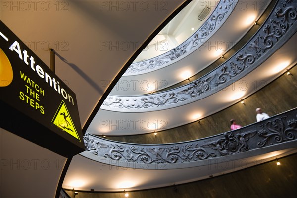 ITALY, Lazio, Rome, Vatican City Museums Tourists descending the Spiral Ramp designed by Giuseppe Momo in 1932 leading from the museums to the street level below seen from below with an illuminated sign warning to watch the steps