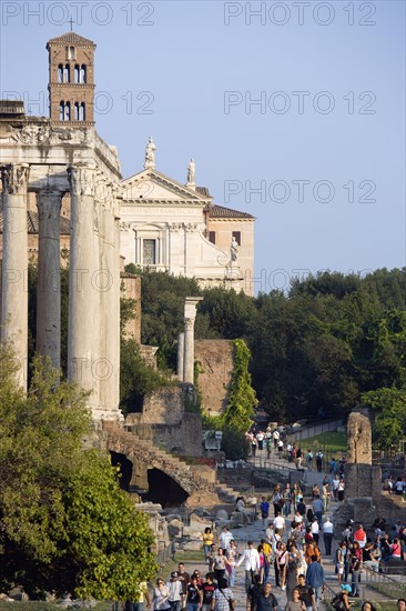 ITALY, Lazio, Rome, Tourists walking past the Temple of Antoninus and Faustina in the Forum with the belltower and facade of the church of Santa Francesca Romana beyond