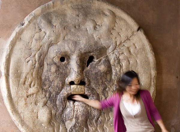 ITALY, Lazio, Rome, Woman with her hand in the jaws of The Bocca della Verita or Mouth of Truth thought to be a medieval drain cover in the portico of the church of Santa Maria in Cosmedin. Tradition had it that the mouth would close over the hands of those who told lies