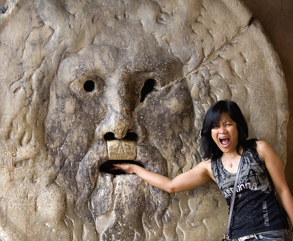 ITALY, Lazio, Rome, Woman with her hand in the jaws of The Bocca della Verita or Mouth of Truth thought to be a medieval drain cover in the portico of the church of Santa Maria in Cosmedin. Tradition had it that the mouth would close over the hands of those who told lies