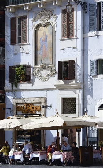 ITALY, Lazio, Rome, People seated outside a restaurant at tables in the shade of umbrellas below a mural of the Virgin Mary on a pale blue building in the Piazza della Rotunda