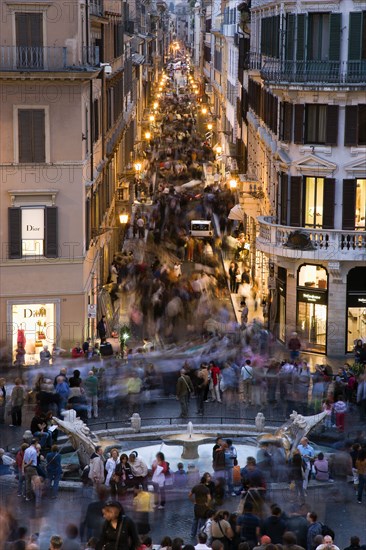 ITALY, Lazio, Rome, The Via dei Condotti the main shopping street busy with people illuminated at night seen from the Spanish Steps with seated tourists and the Fontana della Barcaccia in the foreground