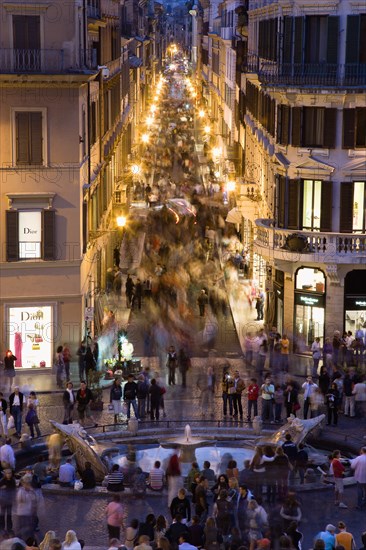 ITALY, Lazio, Rome, The Via dei Condotti the main shopping street busy with people illuminated at night seen from the Spanish Steps with seated tourists and the Fontana della Barcaccia in the foreground