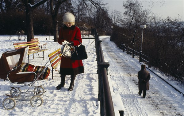 CZECH REPUBLIC, Prague, "Woman stopped to read newspaper in park in the snow.  She wears winter coat, hat and boats with baby in push chair beside her covered with thick blanket."